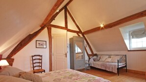 A bedroom with a sloped ceiling, wooden beams, a bed with a floral bedspread, a chair, a wardrobe, and a window with a curtain. - Character house in Crouttes with fireplace and pets allowed (Crouttes)