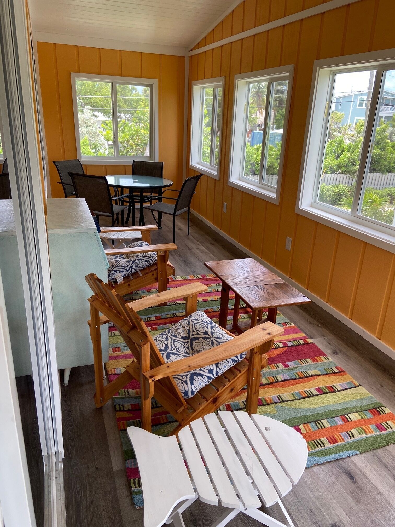 A sunlit room with wooden chairs and a table, large windows, and a colorful rug.