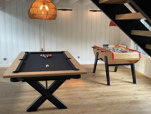 Una mesa de billar con bola blanca, una mesa de fútbol de mesa y un lámpara colgante en una habitación con piso de madera y paredes blancas.