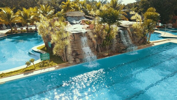 A pool with a waterfall feature surrounded by palm trees and a rocky island. - Ecologic Ville Caldas Vip (Caldas Novas)