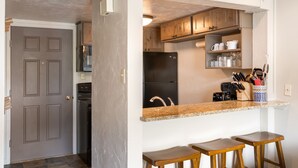 A kitchen with wooden cabinets, a black refrigerator, a granite countertop, and two wooden stools. - Bright Loft Condo | Cathedral Ceilings, Fireplace, and Walk to Slopes! (Steamboat Springs)