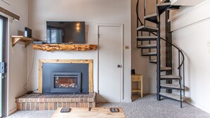 A living room with a fireplace, a TV mounted above it, a wooden coffee table, and a spiral staircase. - Bright Loft Condo | Cathedral Ceilings, Fireplace, and Walk to Slopes! (Steamboat Springs)