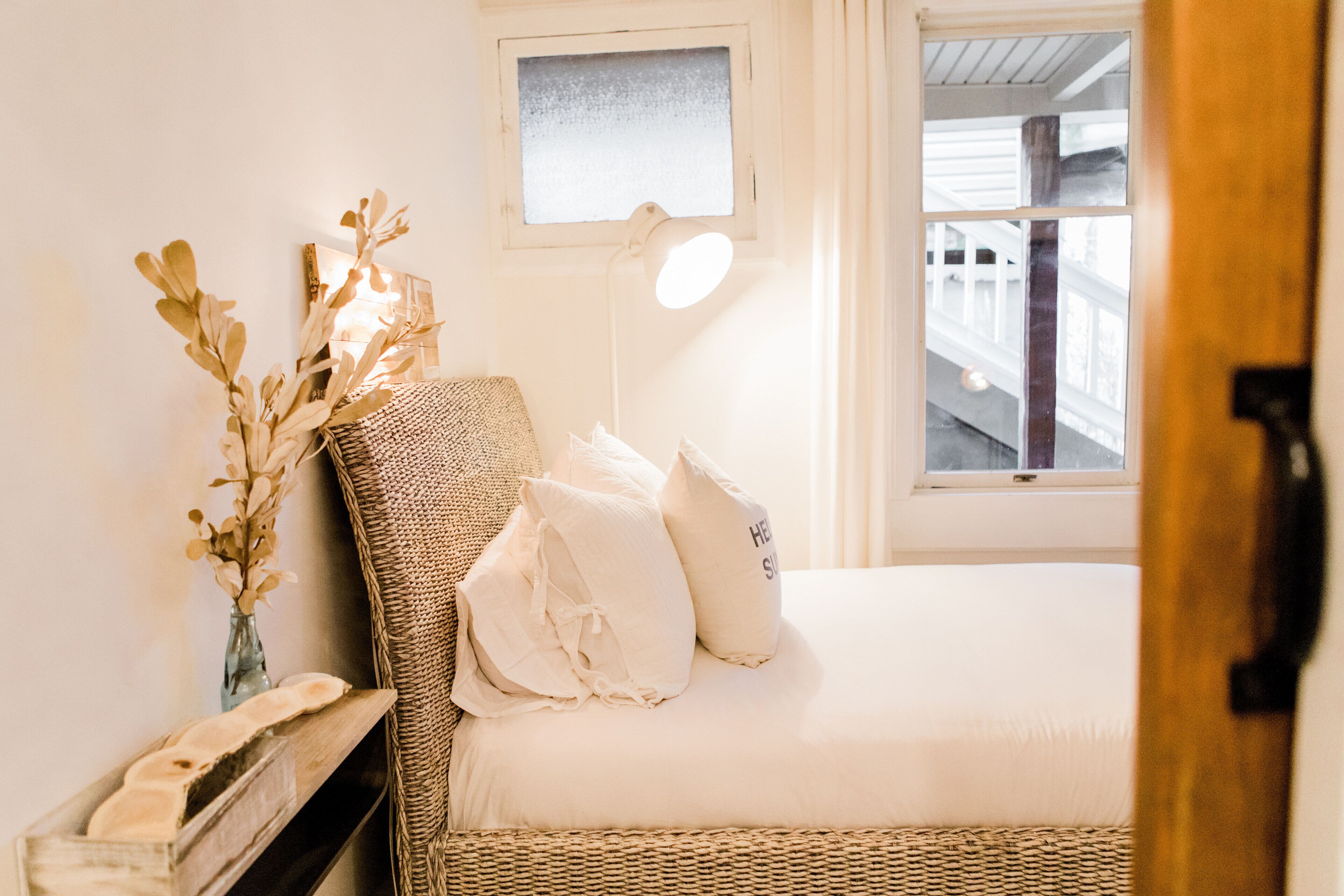 Une chambre à coucher avec un lit, des oreillers, une lampe, un vase contenant des fleurs séchées et une fenêtre.