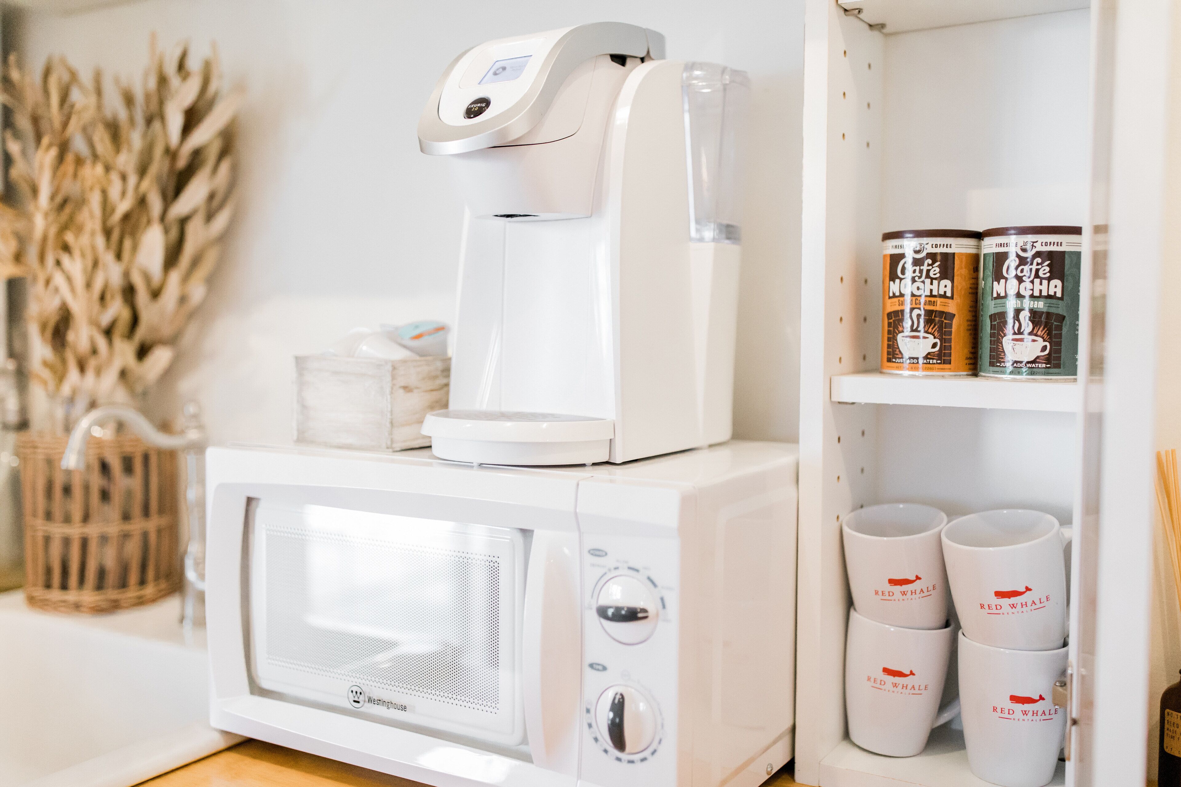 A white microwave with a coffee maker on top, a shelf with two coffee cans, and a basket with a plant.