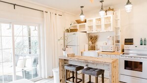 Une cuisine moderne avec des meubles blancs, un îlot central en bois doté de tabourets, un four encastré et une grande fenêtre offrant une vue sur l’extérieur.