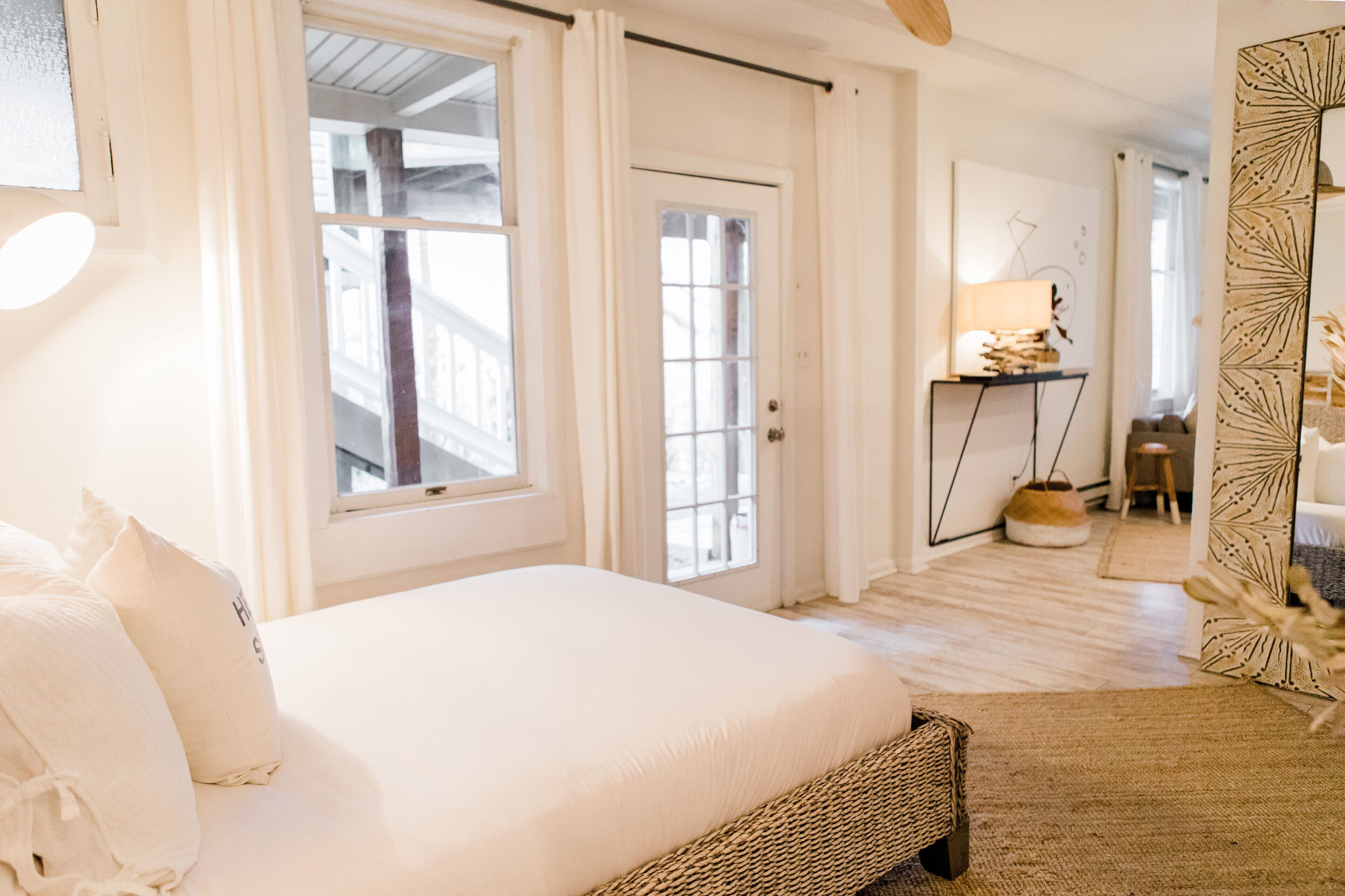 A hotel room with a large bed, a bedside table with a lamp, a mirror with a leaf pattern, and a view of the outside through a window.