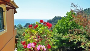 Un balcon orné d’un pot de fleurs rouges et roses, d’où l’on peut admirer un paysage côtier avec des arbres et une vue sur la mer.