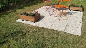 An outdoor seating area with a table and chairs on a tiled surface, surrounded by grass and trees. - Cozy studio near Caen and the sea, landing beaches (Biéville-Beuville)