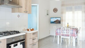 Cocina moderna con estufa a gas, horno y armarios de madera. Cuenta con un comedor con mesa para cuatro personas, un televisor y un reloj en la pared.