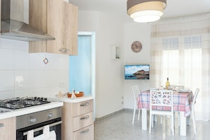 A modern kitchen with a gas stove, oven, and wooden cabinets. There is a dining area with a table set for two, a television, and a window with a view of a coastal town.