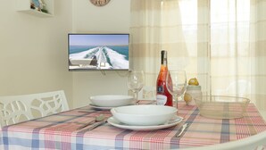 Una mesa de comedor dispuesta con platos blancos, copas de vino y cubiertos, una botella de vino y un pequeño bol. Un televisor que muestra un barco en el agua está montado en la pared.