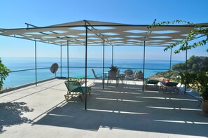 A covered outdoor seating area with a view of the sea, featuring a table and chairs, a potted plant, and a pergola structure.