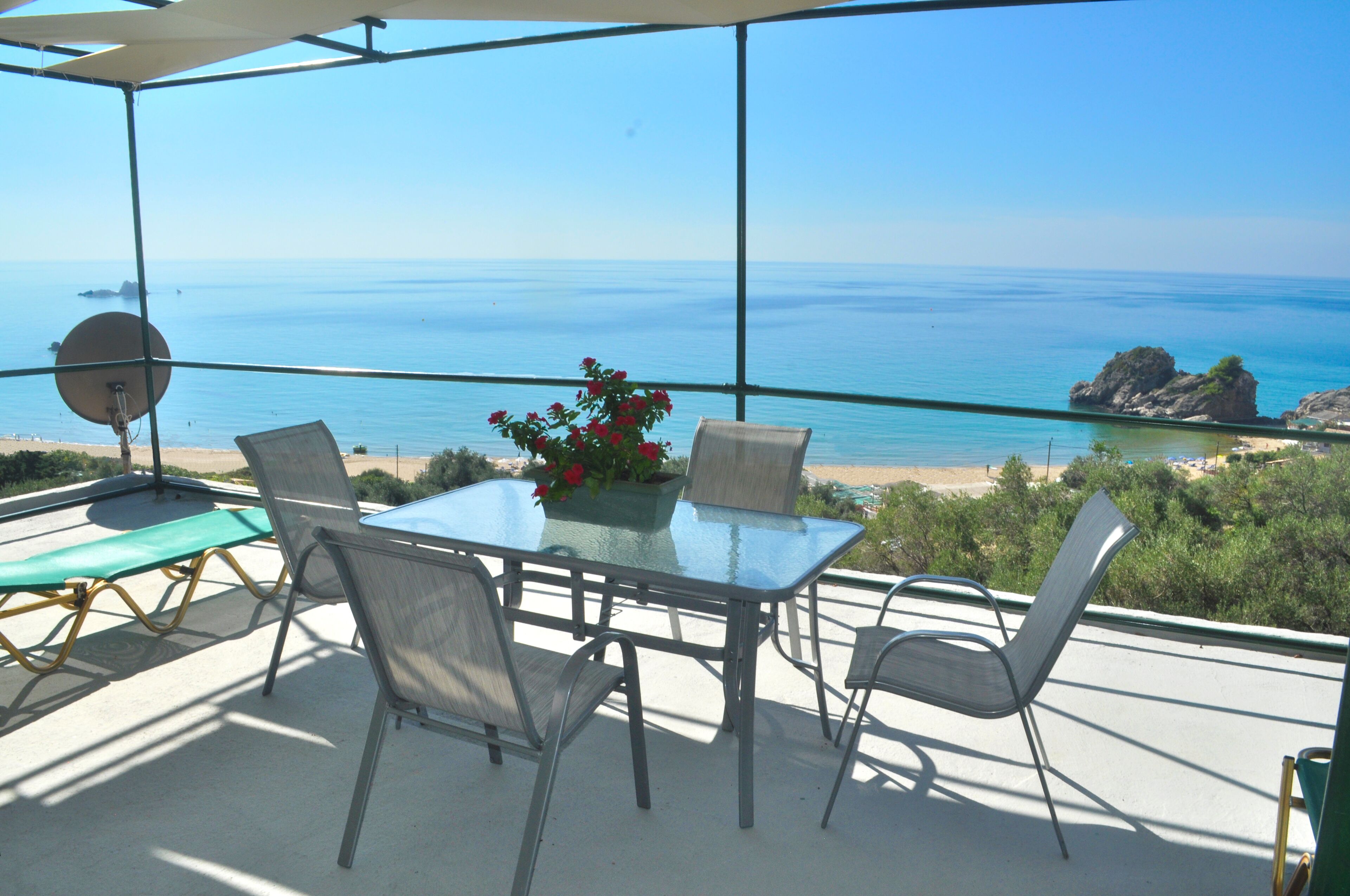 A glass table with chairs and a potted plant on a balcony overlooking a beach and ocean.