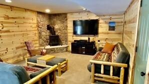A cozy living room with wooden walls, a stone fireplace, a flat-screen TV, and comfortable seating. - Year round fun close to lakes, golf, and ski! (Bellaire)