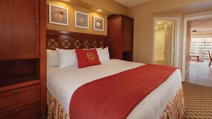 A hotel room with a large bed, wooden headboard, and a red throw blanket. There are framed pictures on the wall and a ceiling fan.