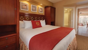 Une chambre d’hôtel avec un grand lit, une tête de lit en bois et un plaid rouge. Des tableaux encadrés ornent le mur et un ventilateur de plafond est installé au plafond.