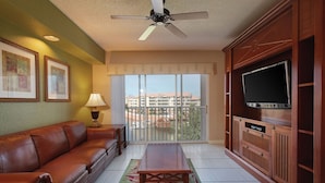 Un salon avec un canapé en cuir, une table basse, un téléviseur posé sur un meuble en bois, un ventilateur de plafond et un balcon offrant une vue sur les immebles.