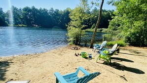 A sandy beach with a lake, two blue lounge chairs, a green chair, and a metal table.