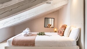 Ett sovrum med ett sluttande tak, en säng med vita sängkläder, en rosa pläd och ett sängbord med en lampa.