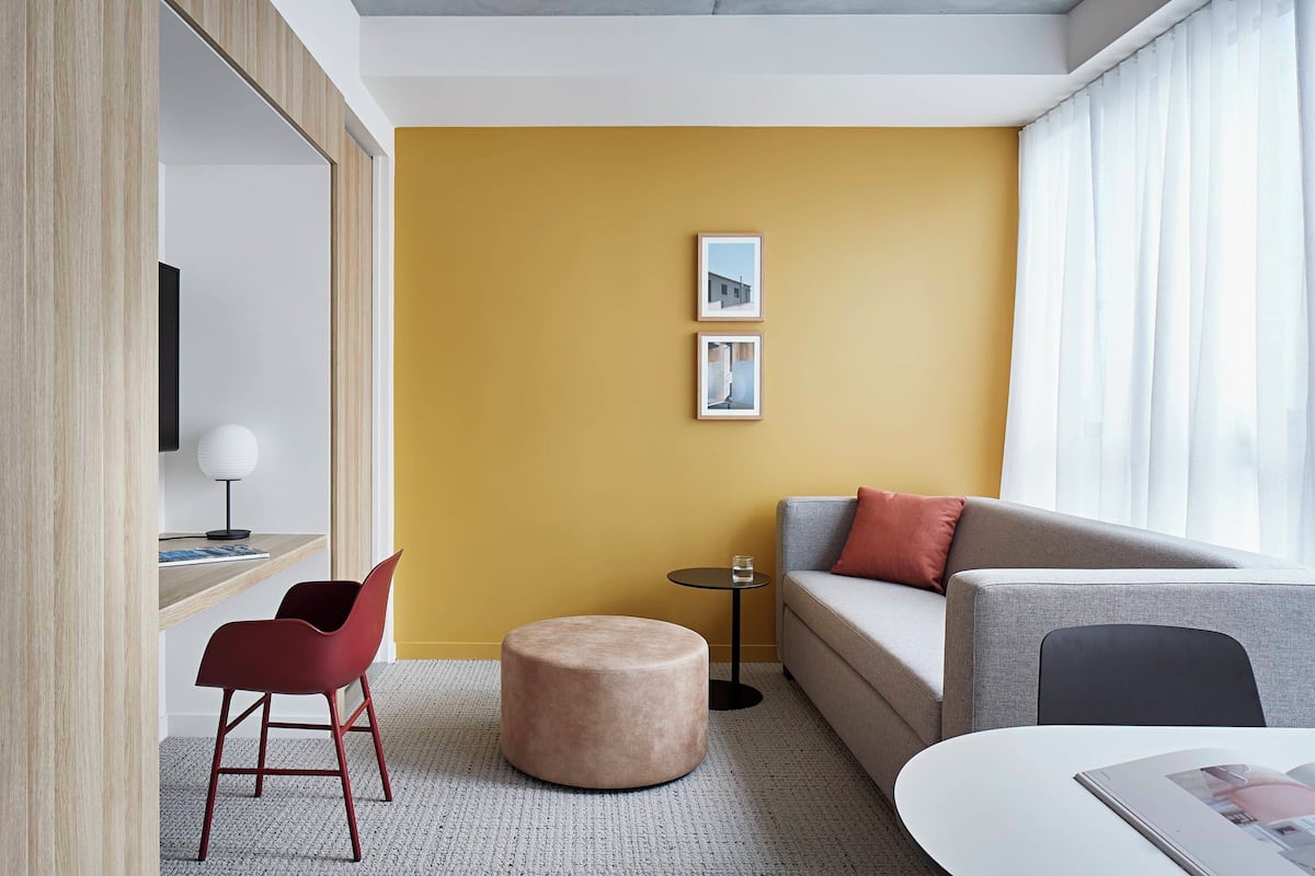 Una habitación de hotel moderna con un sofá, un reposapiés redondo, una mesita auxiliar, un televisor y dos cuadros enmarcados en la pared.