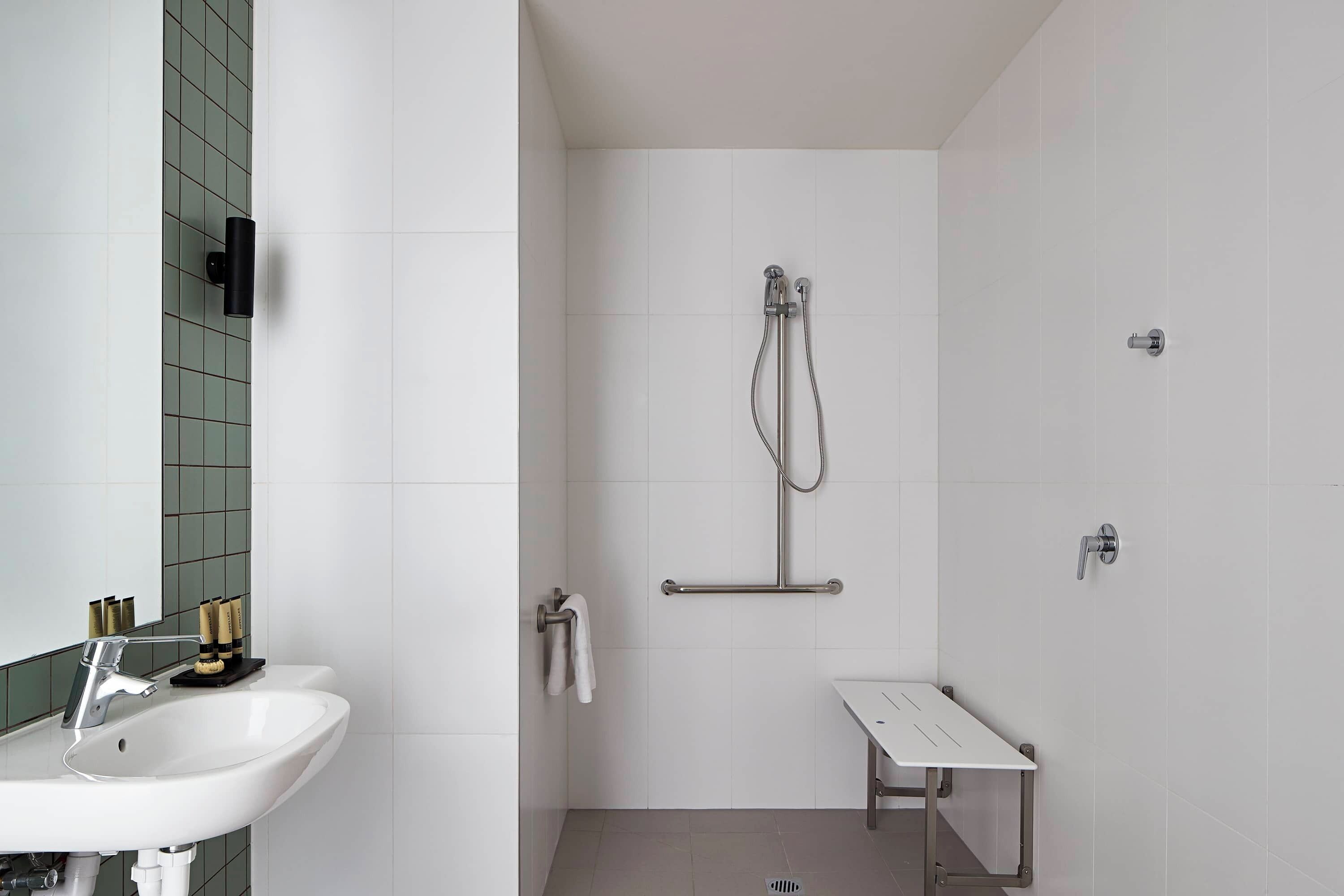 A modern bathroom with a white sink, a handheld showerhead, a towel rack, and a small bench.