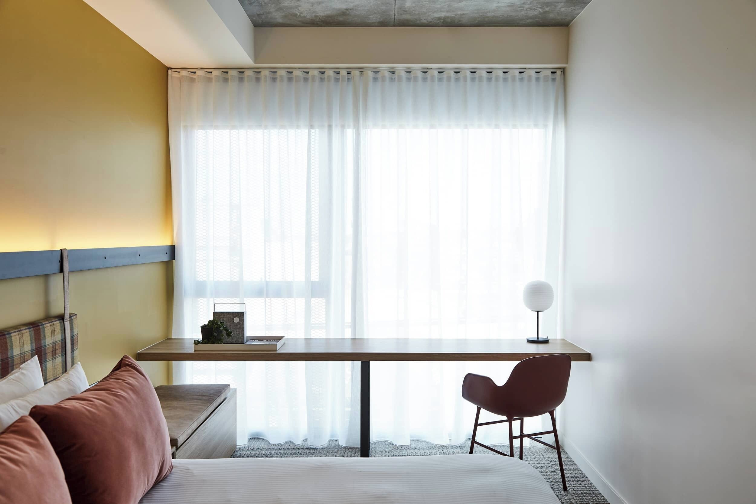 A room with a bed, a desk, a chair, a lamp, and a window with sheer curtains.