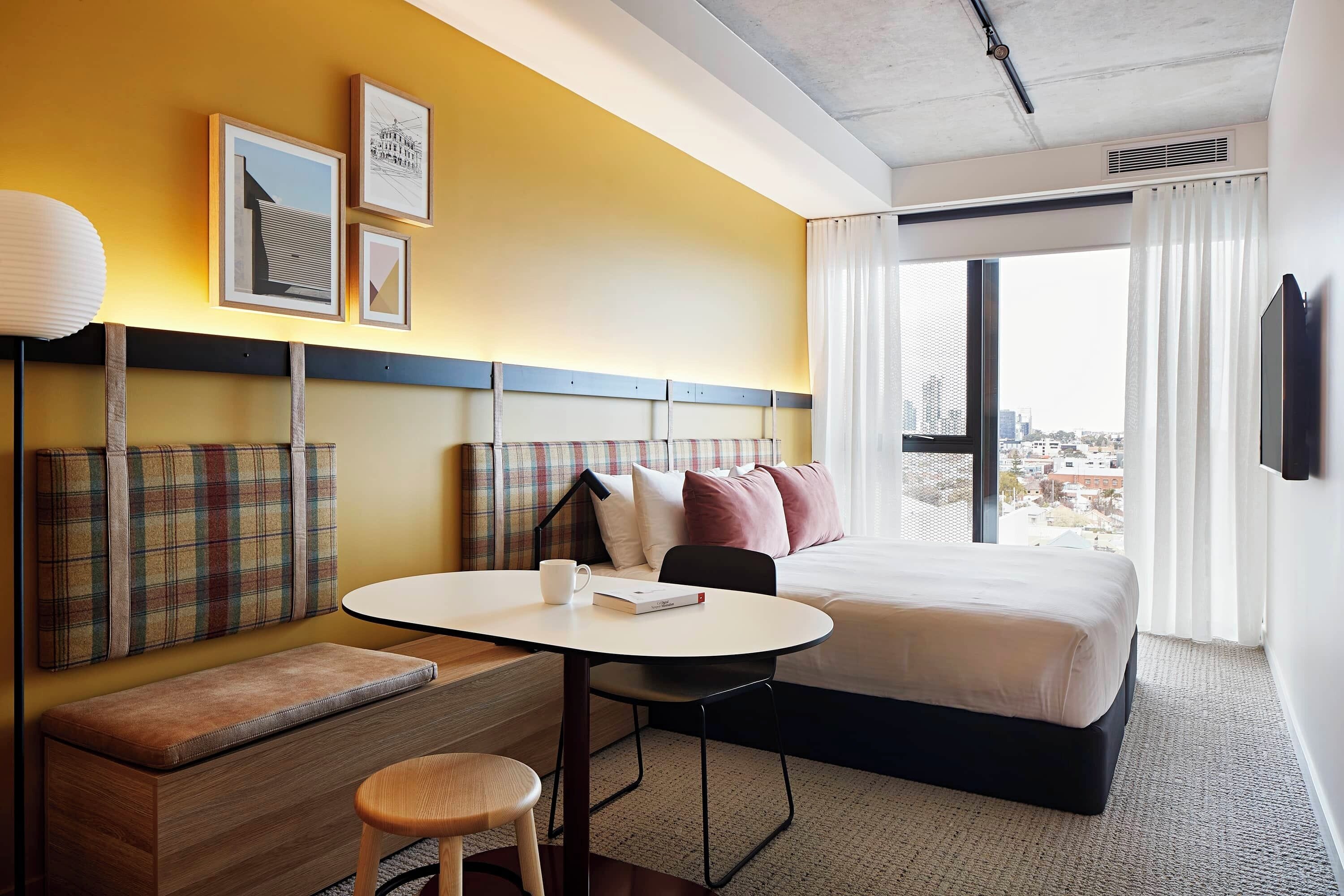 A hotel room with a bed, a desk with a chair, a bench, and a window with a city view.