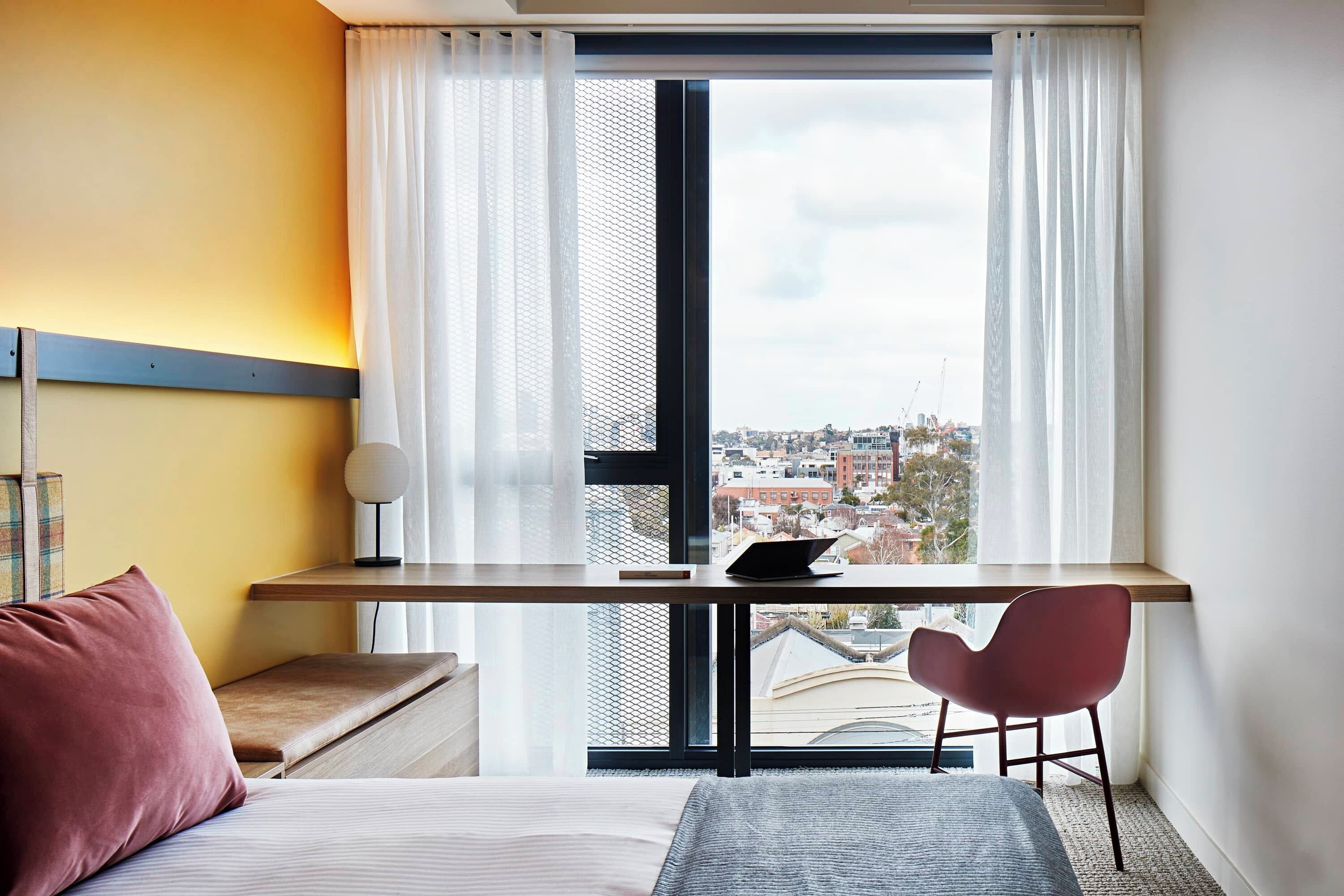 A hotel room with a bed, a desk with a laptop, a chair, and a view of the city through large windows.