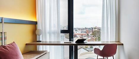 Habitación de hotel con cama, escritorio con computadora portátil, silla y vista de la ciudad a través de amplias ventanas.