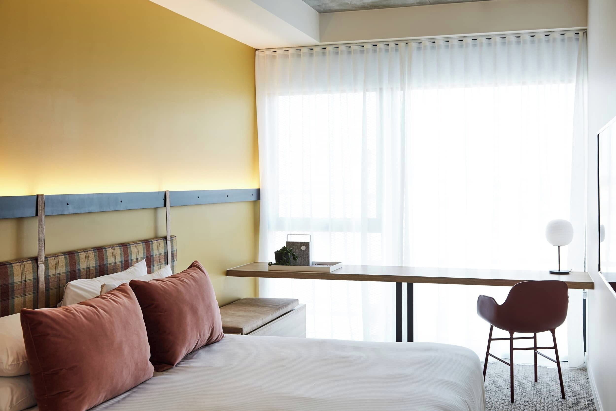 Una habitación de hotel con cama, escritorio, silla y ventana con cortinas translúcidas.