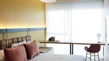 Una habitación de hotel con cama, escritorio, silla y ventana con cortinas translúcidas.