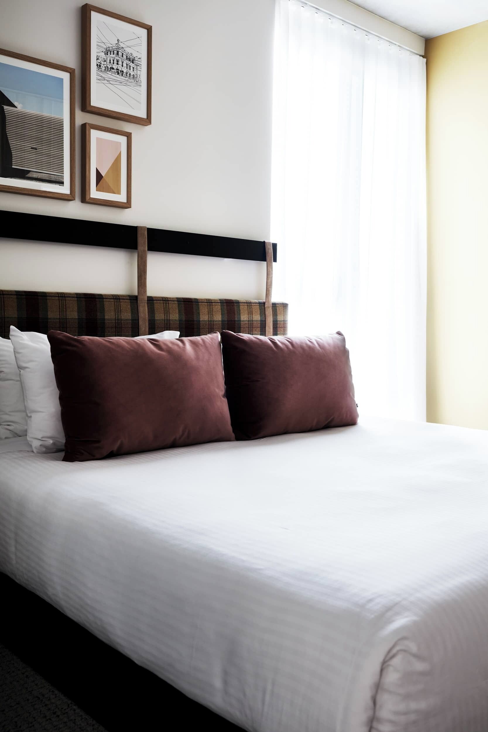 Quarto de hotel com cama, almofadas, cabeceira e quadros na parede.