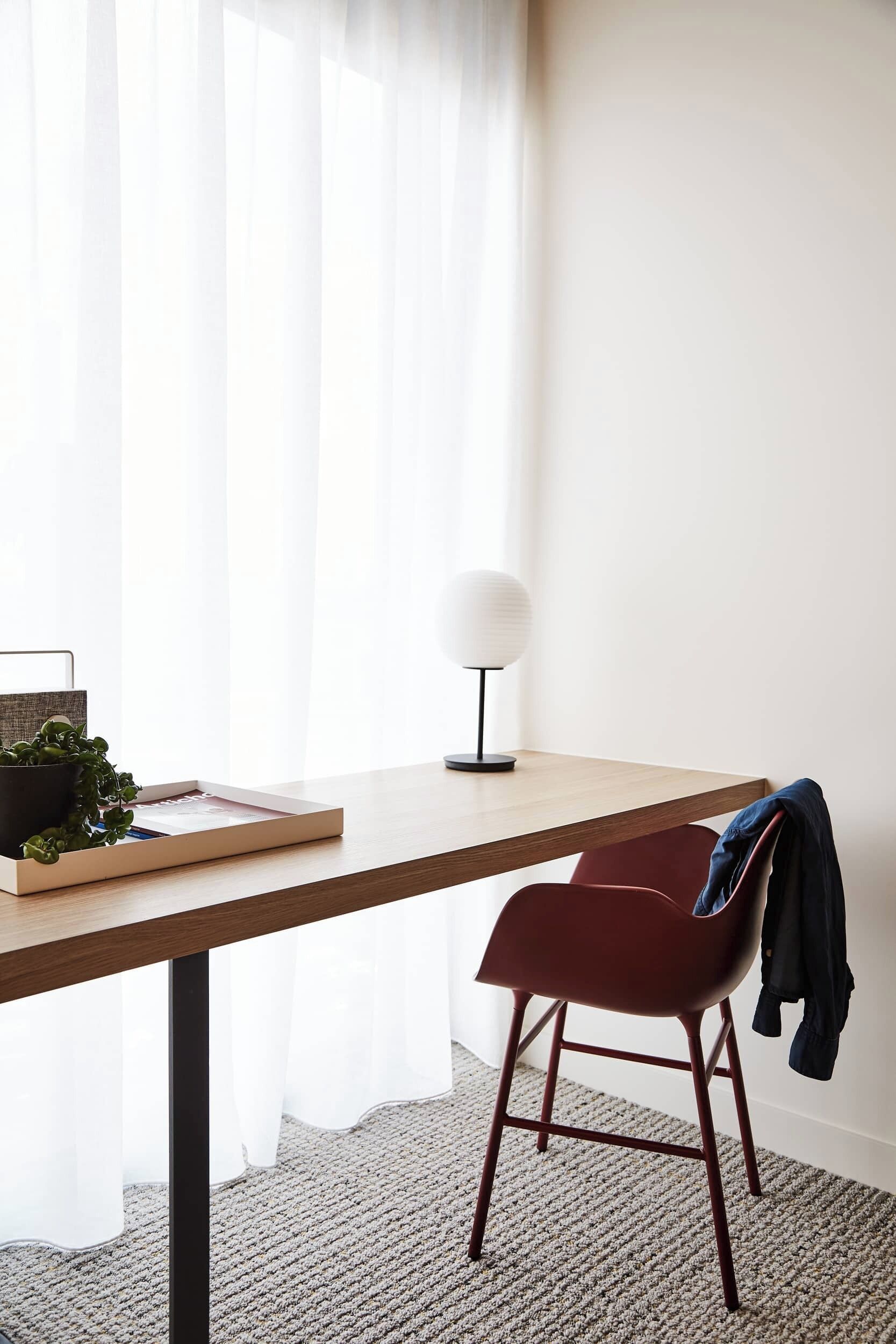 A room with a wooden desk, a red chair, a potted plant, a lamp, and sheer curtains.