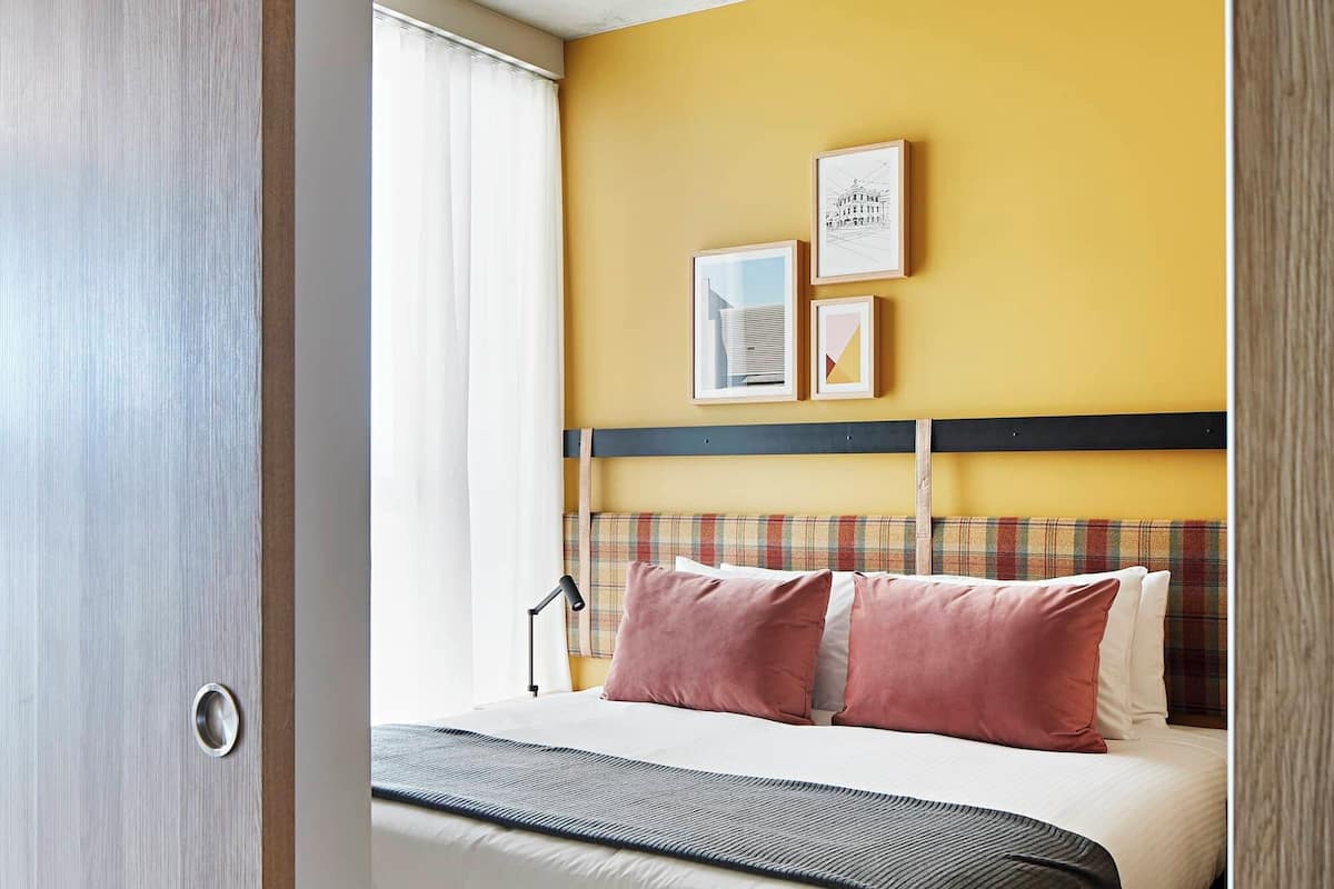 Habitación de hotel con cama, cabecero, ventana con cortinas y pared con cuadros.