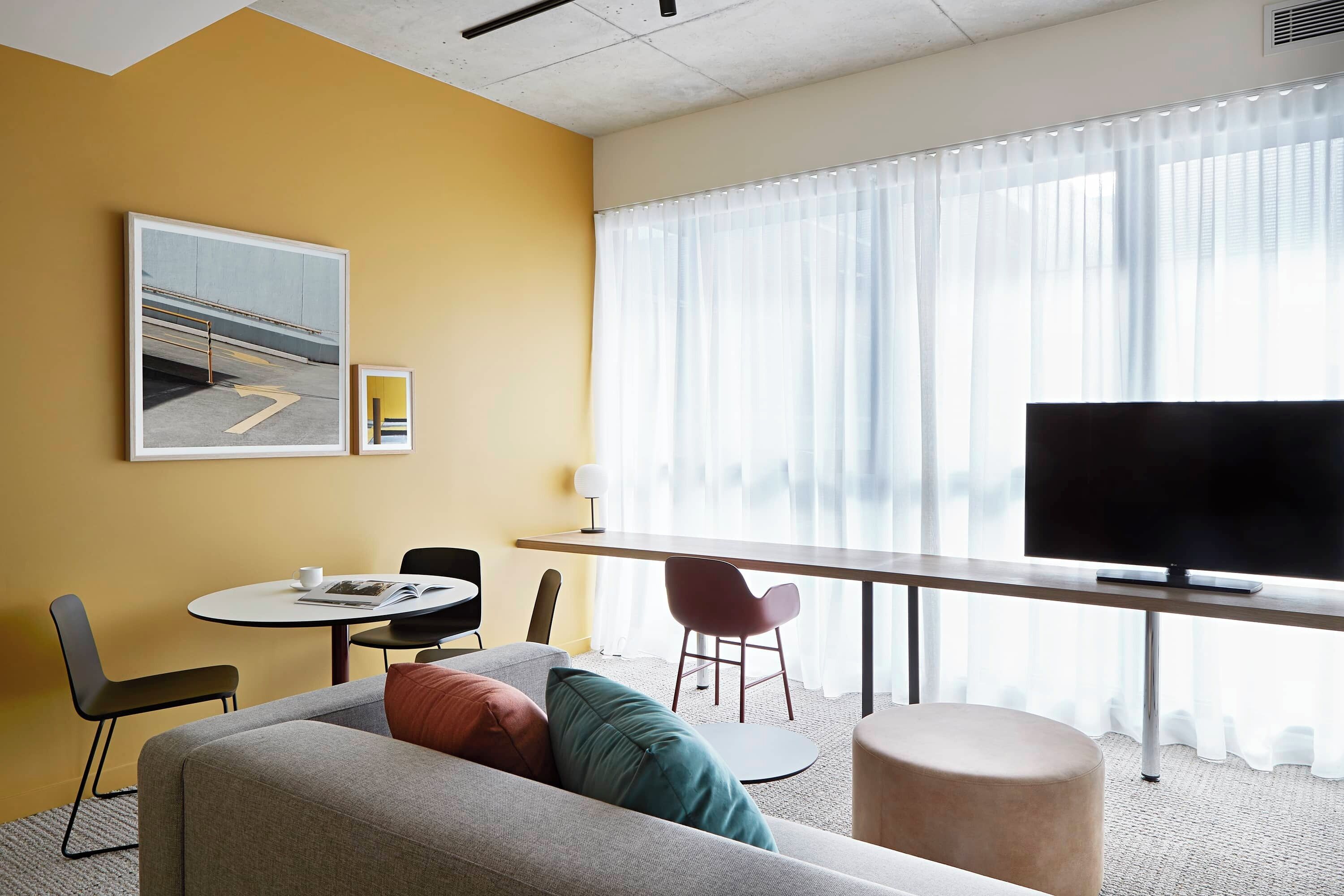 Une chambre d’hôtel moderne équipée d’un canapé, d’une table à manger, d’un téléviseur et d’une grande fenêtre avec des rideaux.