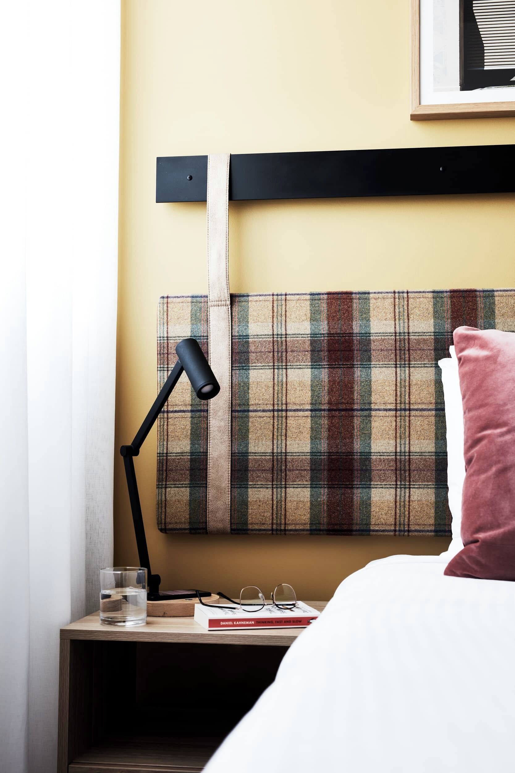 A bed with a plaid headboard, a bedside lamp, a glass of water, and a book on a wooden nightstand.