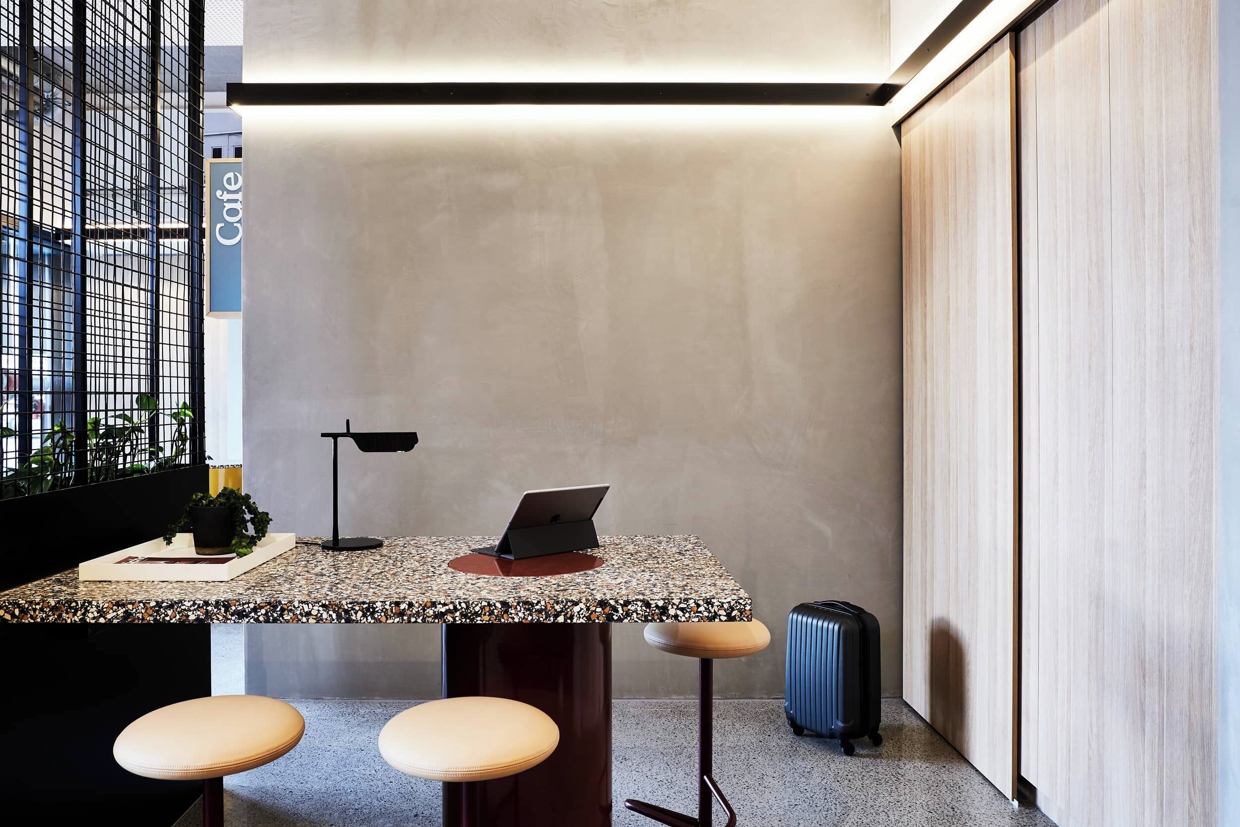 A modern bar area with a laptop on the counter, two stools, a small table with a plant, and a suitcase on the floor.