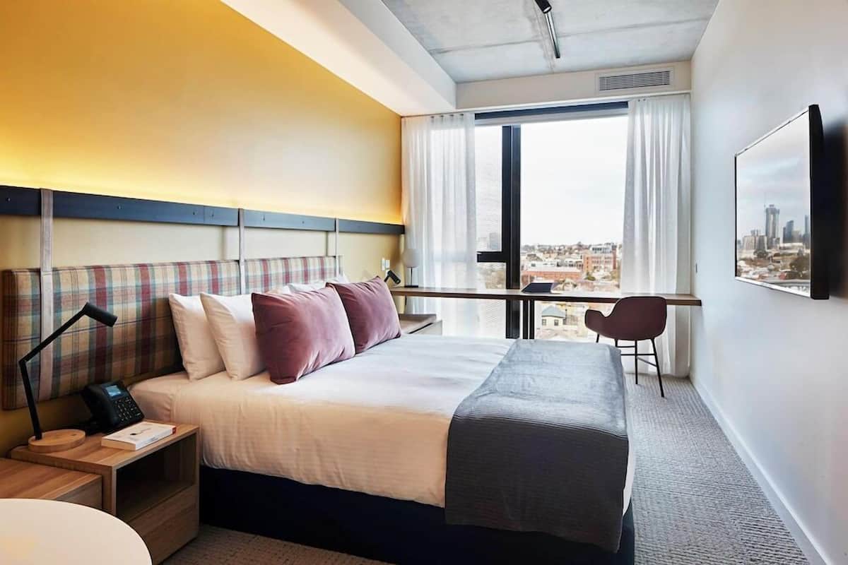 Habitación de hotel con cama, escritorio, silla, vista a la ciudad y televisión montada.
