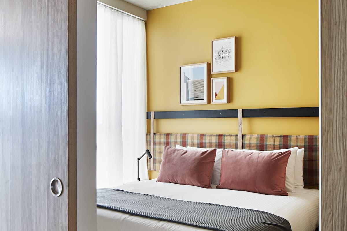 Una habitación de hotel con cama, cabecera, un cuadro colgado en la pared y ventana con cortinas.