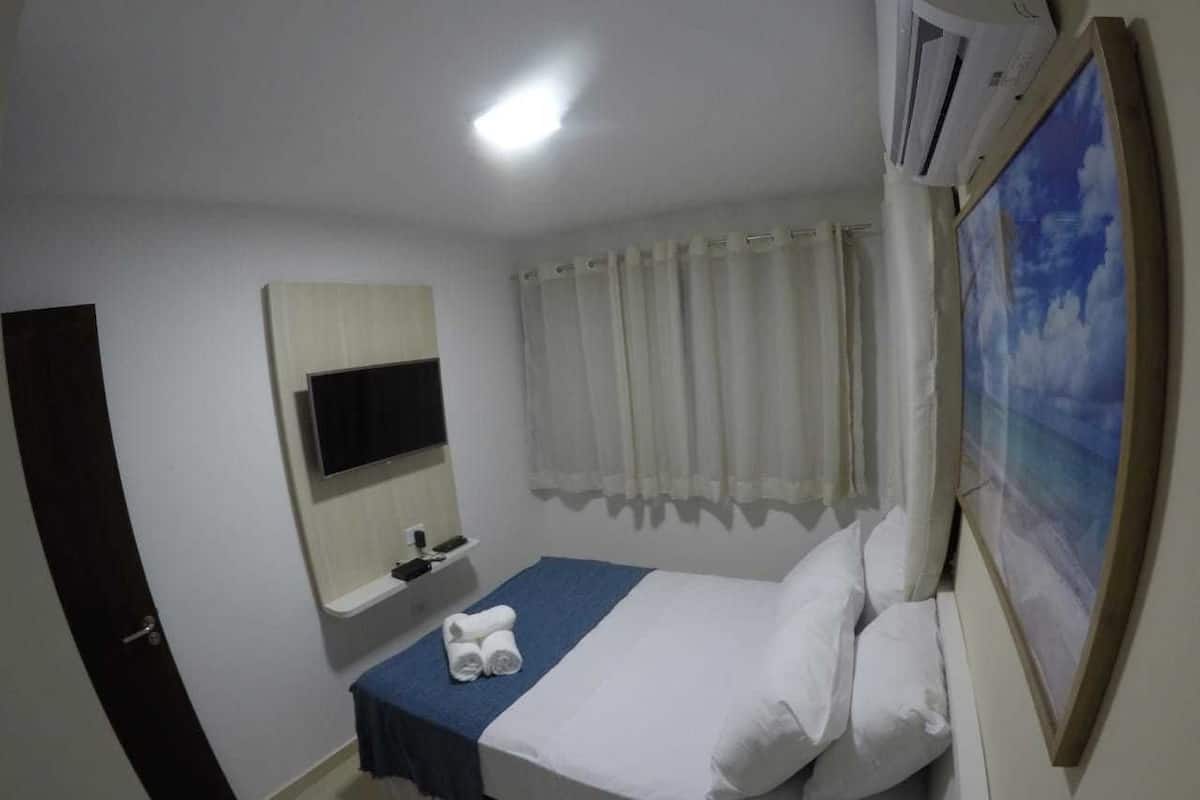 Ein Schlafzimmer mit Bett, einem an der Wand montierten Fernseher, einem Fenster mit Vorhängen und einem Bild an der Wand.