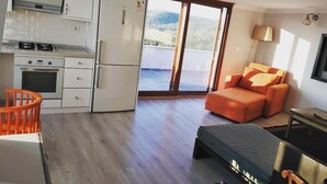 A modern kitchen with white cabinets and appliances, a dining area with a wooden table and orange chairs, and a large sliding glass door leading to a balcony with a view of the mountains. - Tepedeki Ev Apart (Gomec)