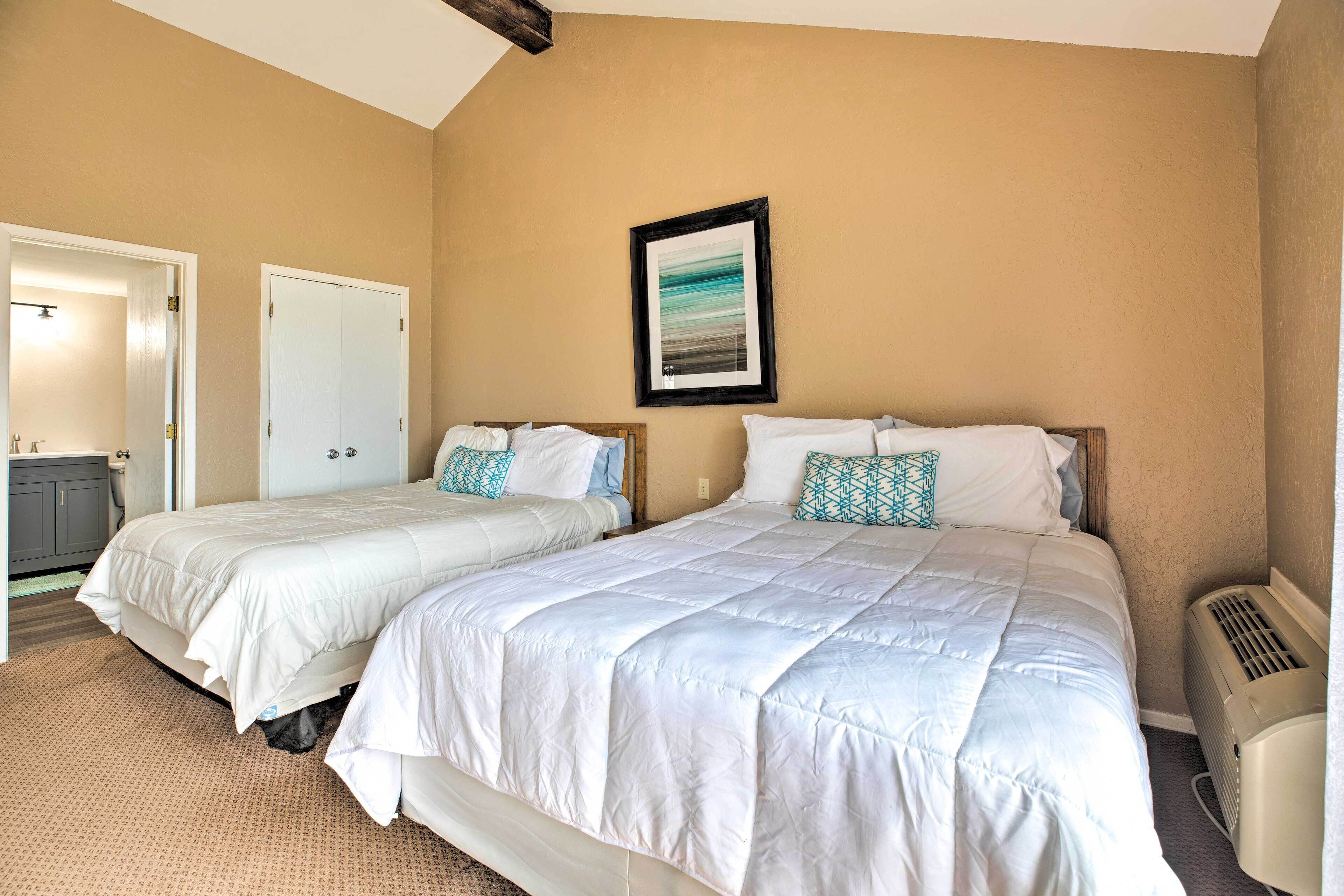 A bedroom with two beds, beige walls, a framed picture, and a ceiling fan.
