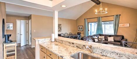 A kitchen with granite countertops, a stainless steel sink, wooden cabinets, a coffee station, and a dining area with a brown leather couch and blue curtains.
