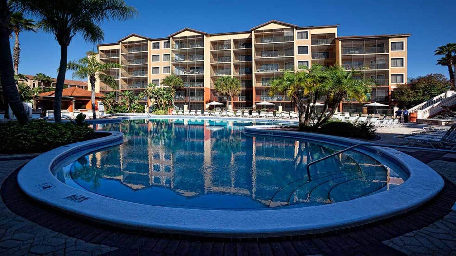 Et hotel med pool, palmer og en klar blå himmel.