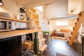 A compact kitchen with a microwave, coffee maker, and wooden shelves. There is a green table and a sofa in the living area. - Oddfellow Inn & Farm (Helena)