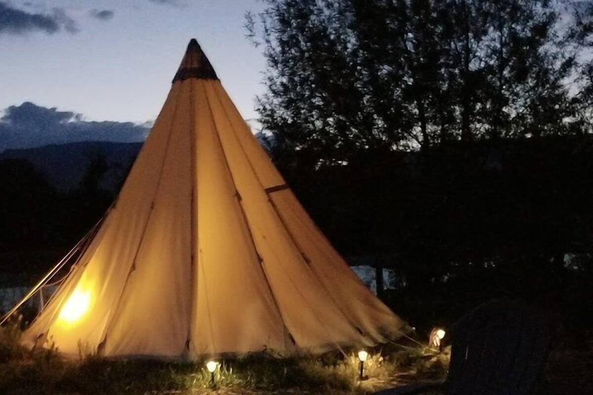 Un'ampia tenda a forma di cono, illuminata al centro, circondata da un'area erbosa e alberi.
