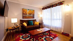 Ein Wohnzimmer mit einem braunen Sofa, zwei Beistelltischen, einem Couchtisch aus Holz, einem farbenfrohen Teppich und einem Gemälde von Bäumen.
