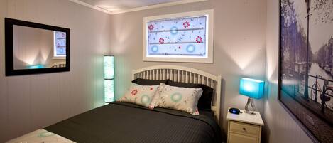Un dormitorio con cama, mesita de noche con lámpara, espejo, ventana con vista a un canal y un cuadro en la pared.