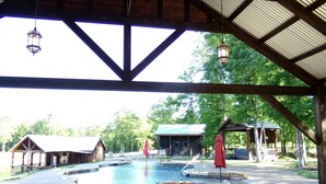 A covered swimming pool area with a wooden deck, a red umbrella, and a view of cabins in the background. - The Bridge House located in an Beautiful RV Resort near Lake Conroe.  (Montgomery)