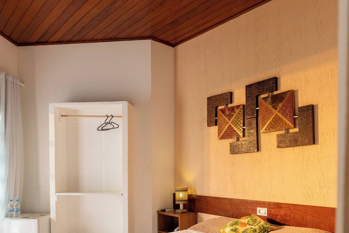 A well-lit bedroom with a wooden ceiling, a bed with a white and beige bedspread, a nightstand with a lamp, and a wall with decorative art pieces.